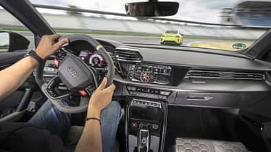 Die Innenraumaufnahme zeigt das Cockpit des Audi RS3 Competition Limited aus der Fahrerperspektive. Sichtbar sind RS‑Sportlenkrad, digitales Kombiinstrument, zentraler Touchscreen und RS‑spezifische Bedienelemente in der Mittelkonsole. Durch die Frontscheibe ist eine Fahrszene auf der Rennstrecke in Hockenheim zu erkennen.