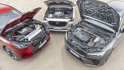Drei moderne SUV mit geöffneten Motorhauben, darunter ein rotes BMW-SUV, ein silbernes Mazda-SUV und ein graues Volvo-SUV. Die Motorenräume sind gut sichtbar, die Fahrzeuge stehen auf einem asphaltierten Platz.