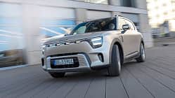 Das Bild zeigt einen Smart #5, ein vollelektrisches Kompakt-SUV, in Fahrt vor moderner urbaner Kulisse. Die Fahrzeugfront und das Nummernschild sind deutlich sichtbar, das Auto befindet sich in Bewegung.