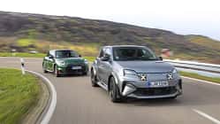 Zwei Elektro-Kompaktsportwagen, ein Alpine A290GTS und ein Mini John Cooper Works E, fahren auf einer kurvigen Landstraße. Beide Fahrzeuge sind in Bewegung, der Alpine vorne in mattem Grau, der Mini dahinter in Dunkelgrün. Im Hintergrund sind hügelige Landschaft und herbstliche Vegetation sichtbar.