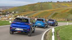 Drei kompakte SUV-Modelle – Opel Mokka, Ford Puma und VW Taigo – fahren in einer Gruppe auf einer kurvigen Landstraße durch eine hügelige Landschaft mit Weinbergen.