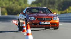 Mercedes Benz SLK 230 Kompressor fährt auf Teststrecke durch Slalom mit Pylonen. Cabrio in orange, Frontansicht, Fahrer sichtbar.
