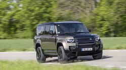 Land Rover Defender 130 D250, SUV, Geländewagen, Allradantrieb, Offroad, Land Rover, Defender, Straßenfahrt, Automobil, Fahrzeug Land Rover Defender 130 D250 fährt auf einer Landstraße, seitliche Frontansicht, grüne Natur im Hintergrund.