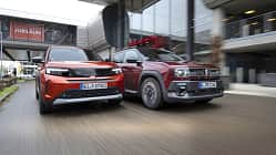 Opel Frontera Electric und Renault 4 E-Tech 150 fahren nebeneinander auf einer Straße vor moderner Architektur.
