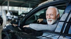 07/2026, Senior im Auto Ein älterer Mann mit weißem Bart am Steuer eines Pkw schaut lächelnd aus einem geöffneten Autofenster