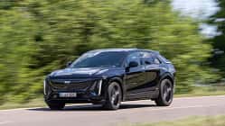 Cadillac Lyriq-V fährt auf einer Landstraße, Seitenansicht, schwarze Lackierung, grüne Vegetation im Hintergrund.