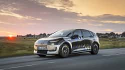 Audi A2 e-tron, Erlkönig, Elektroauto, Audi, Kompaktwagen, Tarnfahrzeug, Prototyp, Elektromobilität, Straßenfahrt, Sonnenuntergang Audi A2 e-tron Erlkönig (Tarnfahrzeug) fährt auf Landstraße bei Sonnenuntergang. Das Fahrzeug trägt eine auffällige Tarnfolierung, im Hintergrund ist eine ländliche Szenerie mit Häusern und Wiesen zu sehen.