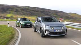 Zwei Elektro-Kompaktsportwagen, ein Alpine A290GTS und ein Mini John Cooper Works E, fahren auf einer kurvigen Landstraße. Beide Fahrzeuge sind in Bewegung, der Alpine vorne in mattem Grau, der Mini dahinter in Dunkelgrün. Im Hintergrund sind hügelige Landschaft und herbstliche Vegetation sichtbar.
