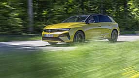 Opel Astra PHEV in Gelb fährt auf einer Landstraße durch grüne, bewaldete Umgebung. Das Fahrzeug ist im Profil zu sehen, mit sichtbarem Fahrer und dynamischer Bewegungsunschärfe im Vordergrund.