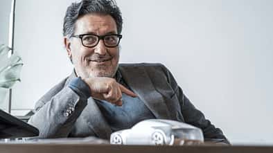 Porträtaufnahme einer Person im Sakko an einem Schreibtisch mit einem Automodell im Vordergrund.