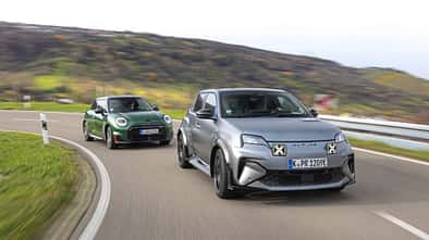 Zwei Elektro-Kompaktsportwagen, ein Alpine A290GTS und ein Mini John Cooper Works E, fahren auf einer kurvigen Landstraße. Beide Fahrzeuge sind in Bewegung, der Alpine vorne in mattem Grau, der Mini dahinter in Dunkelgrün. Im Hintergrund sind hügelige Landschaft und herbstliche Vegetation sichtbar.