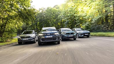 Vier kompakte SUV-Modelle – Dacia Bigster Hybrid, Opel Frontera Hybrid, Hyundai Kona 1.6-GDI HEV und Skoda Karoq 1.5 TSI – fahren nebeneinander auf einer Straße durch eine bewaldete Umgebung.