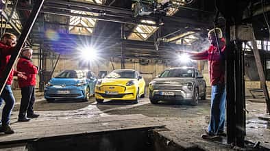 Drei Elektroautos – Ford Puma Gen-E, Kia EV3 und VW ID.3 – stehen nebeneinander in einer alten Industriehalle. Mehrere Personen in roten Jacken stehen um die Fahrzeuge, die Szene ist beleuchtet und vermittelt eine Test- oder Vergleichssituation.