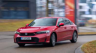 Honda Civic e-HEV in Rot fährt auf einer städtischen Straße, modernes Umfeld, Fahrzeug in Bewegung.