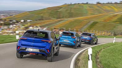 Drei kompakte SUV-Modelle – Opel Mokka, Ford Puma und VW Taigo – fahren in einer Gruppe auf einer kurvigen Landstraße durch eine hügelige Landschaft mit Weinbergen.