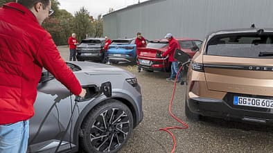 Mehrere Elektro-SUVs (Hyundai IONIQ 5, VW ID.4 Pro, Opel Grandland, Kia EV6, Renault Scenic E-Tech Electric) werden von Personen an Ladestationen geladen. Die Szene zeigt einen Vergleichstest moderner Elektrofahrzeuge.