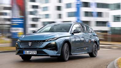 Peugeot e-308 SW 156 PS fährt auf einer städtischen Straße vor modernen Gebäuden.