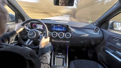 Innenraumaufnahme eines Mercedes GLA250 4Matic während der Fahrt, Blick über die Schulter des Fahrers auf das Cockpit und die Straße.