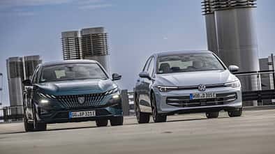Zwei Fahrzeuge, ein Peugeot 308 PHEV und ein VW Golf eHybrid, nebeneinander auf einem Parkdeck in der Stadt.