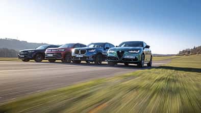 Vier Mittelklasse-SUVs – Mercedes GLC 220d 4Matic, BMW X3 xDrive 20d, Alfa Romeo Stelvio 2.0 Diesel und VW Tiguan 2.0 TDI – fahren nebeneinander auf einer Landstraße.