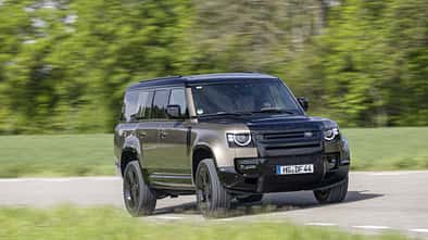 Land Rover Defender 130 D250 fährt auf einer Landstraße, seitliche Frontansicht, grüne Natur im Hintergrund.