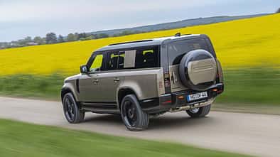 Land Rover Defender 130 D250 fährt auf einer Landstraße an einem blühenden Rapsfeld vorbei.