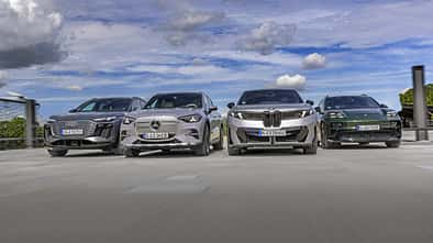 Vier Elektro-SUVs – Audi Q6 SUV e-tron quattro, BMW iX3 50 xDrive, Mercedes GLC 400 4matic EQ und Porsche Macan 4S – nebeneinander auf einer offenen Fläche, fotografiert aus niedriger Perspektive, mit Himmel und Wolken im Hintergrund.