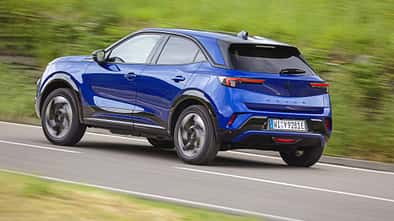 Opel Mokka in blauer Lackierung fährt auf einer Landstraße, fotografiert von schräg hinten.