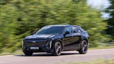 Cadillac Lyriq-V fährt auf einer Landstraße, Seitenansicht, schwarze Lackierung, grüne Vegetation im Hintergrund.