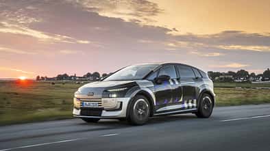 Audi A2 e-tron Erlkönig (Tarnfahrzeug) fährt auf Landstraße bei Sonnenuntergang. Das Fahrzeug trägt eine auffällige Tarnfolierung, im Hintergrund ist eine ländliche Szenerie mit Häusern und Wiesen zu sehen.