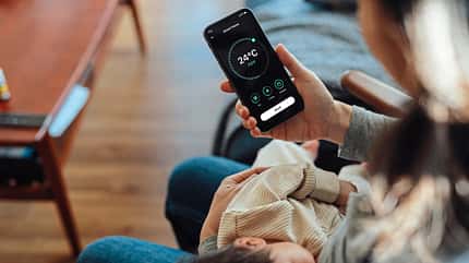 EON Young woman using smart phone to adjust smart thermostat for air-conditioning at home while holding her baby to sleep