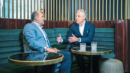 Luca de Meo und Redakteur Stefan Cerchez sitzen in einer Lounge auf einem grünen Sofa und führen ein Gespräch. Auf den Tischen vor ihnen stehen Gläser mit Wasser. Die Szene wirkt wie ein Interview oder Geschäftsgespräch in gehobenem Ambiente.