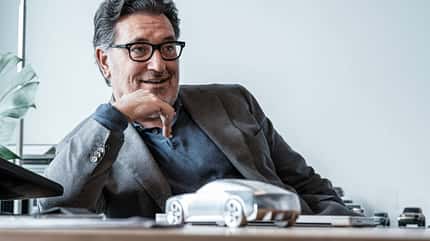 Porträt eines Mannes mit Brille und Bart, der entspannt an einem Schreibtisch sitzt. Im Vordergrund steht ein silbernes Automodell, im Hintergrund sind weitere Modellautos zu sehen.