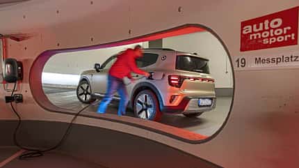 Das Bild zeigt einen Smart #5 beim Ladevorgang an einer Ladestation in einer Tiefgarage. Ein Mann schließt das Ladekabel an das Fahrzeug an. Die Szene ist durch ein rundes Fenster in der Wand fotografiert, im Vordergrund ist ein Schild mit 'auto motor und sport' und 'Messplatz 19' sichtbar.