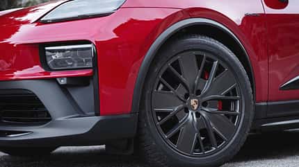 Detailaufnahme eines Vorderrads an einem roten Porsche-Elektrofahrzeug. Das Bild zeigt die Felge mit Porsche-Logo, den Reifen und einen Teil des Frontkotflügels mit Scheinwerfer.