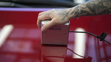 Sonax Lackversiegelung, KRÜSS Mobile Surface Analyzer, Oberflächenmessgerät, Autolack, Lackanalyse, Messgerät, Hand, Tätowierung, SONAX, Fahrzeugpflege, Glanzmessung Eine tätowierte Hand hält ein KRÜSS Mobile Surface Analyzer Messgerät auf einer glänzenden roten Oberfläche, vermutlich ein Autolack. Das Gerät ist per Kabel verbunden und reflektiert auf der Oberfläche.