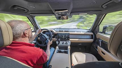 Land Rover Defender 130 D250, SUV-Innenraum, Cockpit, Fahrerperspektive, Allradfahrzeug, Armaturenbrett, Infotainmentsystem, Geländewagen, 2024, Straßenfahrt Innenraumaufnahme eines Land Rover Defender 130 D250 während der Fahrt, Blick über die Schulter des Fahrers auf das Cockpit und die Straße.