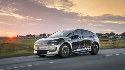Audi A2 e-tron, Erlkönig, Elektroauto, Audi, Kompaktwagen, Tarnfahrzeug, Prototyp, Elektromobilität, Straßenfahrt, Sonnenuntergang Audi A2 e-tron Erlkönig (Tarnfahrzeug) fährt auf Landstraße bei Sonnenuntergang. Das Fahrzeug trägt eine auffällige Tarnfolierung, im Hintergrund ist eine ländliche Szenerie mit Häusern und Wiesen zu sehen.