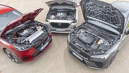 Drei moderne SUV mit geöffneten Motorhauben, darunter ein rotes BMW-SUV, ein silbernes Mazda-SUV und ein graues Volvo-SUV. Die Motorenräume sind gut sichtbar, die Fahrzeuge stehen auf einem asphaltierten Platz.