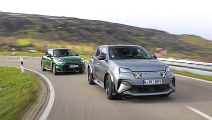 Zwei Elektro-Kompaktsportwagen, ein Alpine A290GTS und ein Mini John Cooper Works E, fahren auf einer kurvigen Landstraße. Beide Fahrzeuge sind in Bewegung, der Alpine vorne in mattem Grau, der Mini dahinter in Dunkelgrün. Im Hintergrund sind hügelige Landschaft und herbstliche Vegetation sichtbar.