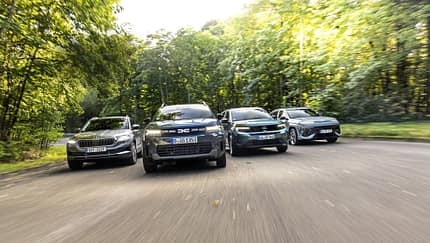 Vier kompakte SUV-Modelle – Dacia Bigster Hybrid, Opel Frontera Hybrid, Hyundai Kona 1.6-GDI HEV und Skoda Karoq 1.5 TSI – fahren nebeneinander auf einer Straße durch eine bewaldete Umgebung.