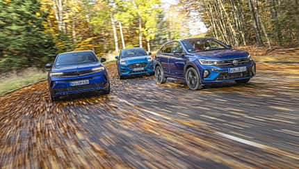 Drei kompakte SUV-Modelle – VW Taigo 1.5 TSI, Opel Mokka Hybrid und Ford Puma 1.0 EcoBoost – fahren nebeneinander auf einer herbstlichen Landstraße.