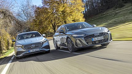 Audi A6 TFSI quattro und Mercedes E450 4matic fahren nebeneinander auf einer Landstraße im Herbst.