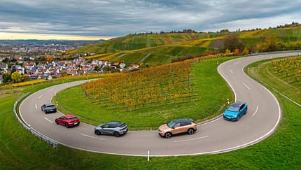 Fünf Elektro-SUVs (Hyundai IONIQ 5, VW ID.4 Pro, Opel Grandland, Kia EV6, Renault Scenic E-Tech Electric) fahren auf einer kurvigen Landstraße durch Weinberge. Die Aufnahme stammt von einer Drohne und zeigt die Fahrzeuge in einer engen Serpentine mit Blick auf eine Ortschaft und herbstliche Weinberge.