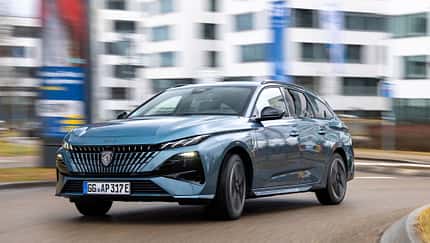Peugeot e-308 SW 156 PS fährt auf einer städtischen Straße vor modernen Gebäuden.