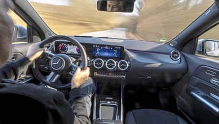 Innenraumaufnahme eines Mercedes GLA250 4Matic während der Fahrt, Blick über die Schulter des Fahrers auf das Cockpit und die Straße.