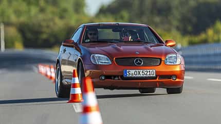 Mercedes Benz SLK 230 Kompressor fährt auf Teststrecke durch Slalom mit Pylonen. Cabrio in orange, Frontansicht, Fahrer sichtbar.