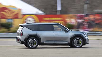 Kia EV9 GT (6 Sitzer) fährt in der Stadt, Seitenansicht, silberner Elektro-SUV vor urbanem Hintergrund.