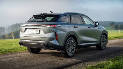 Das Bild zeigt einen OMODA 9 SUV in silberner Lackierung, fotografiert von schräg hinten im Außenbereich auf einer Landstraße. Das Fahrzeug steht auf einer asphaltierten Straße, im Hintergrund ist eine grüne Wiese und eine Baumreihe zu sehen. Die Aufnahme hebt das moderne Design und die markanten LED-Rückleuchten des OMODA 9 hervor.