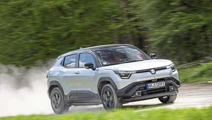 Suzuki e-Vitara eAxle SUV fährt dynamisch auf einer Landstraße, im Hintergrund grüne Vegetation.
